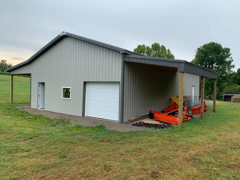 Light gray metal barn with a roll-up door, a side door, and a covered side porch with agricultural equipment underneath.