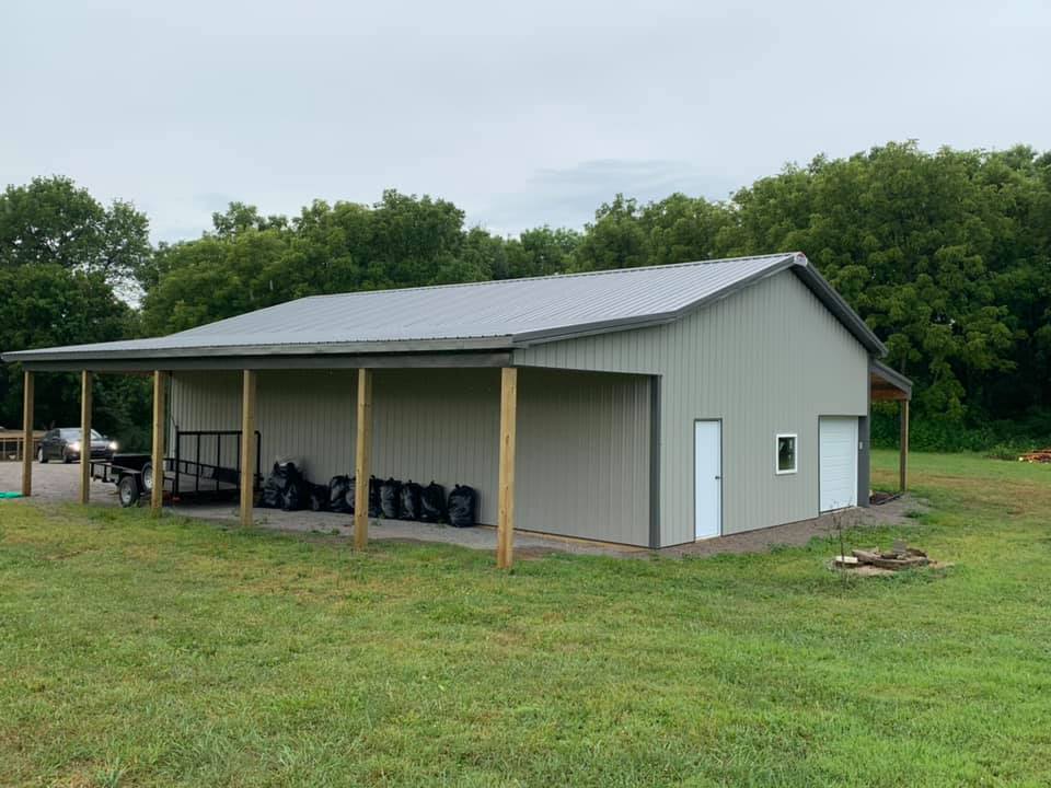 A light-grey metal storage building with a side porch, parked trailer, and surrounding green grass and trees.