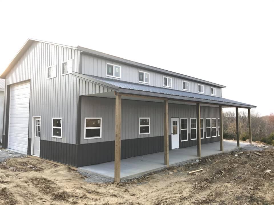 A two-tone gray metal barn with a front porch, a tall rolling garage door, and multiple windows on a dirt lot.