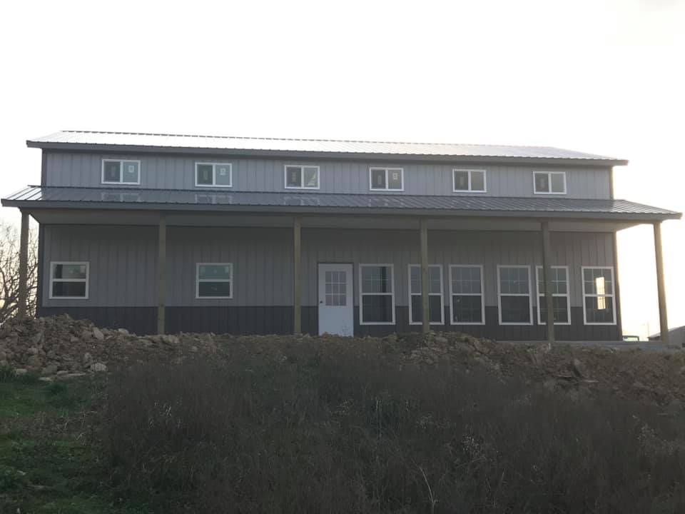 A two-story grey metal building with a covered porch, numerous windows, and a tin roof, set against a bright, clear sky.