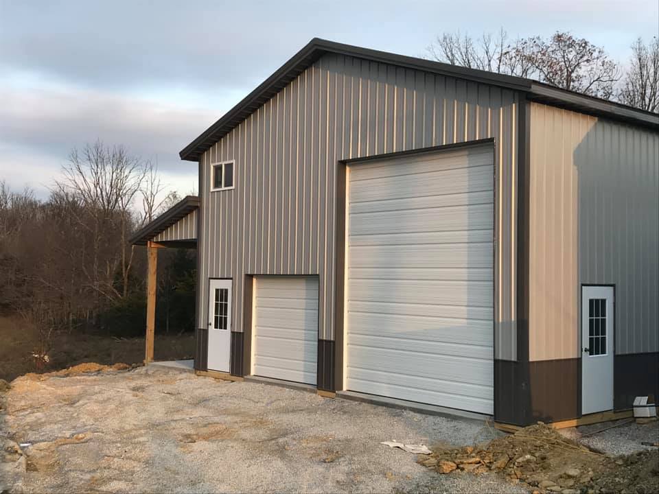 A light gray metal barn with white garage doors and side entry doors, set in a rural area with gravel in the foreground.
