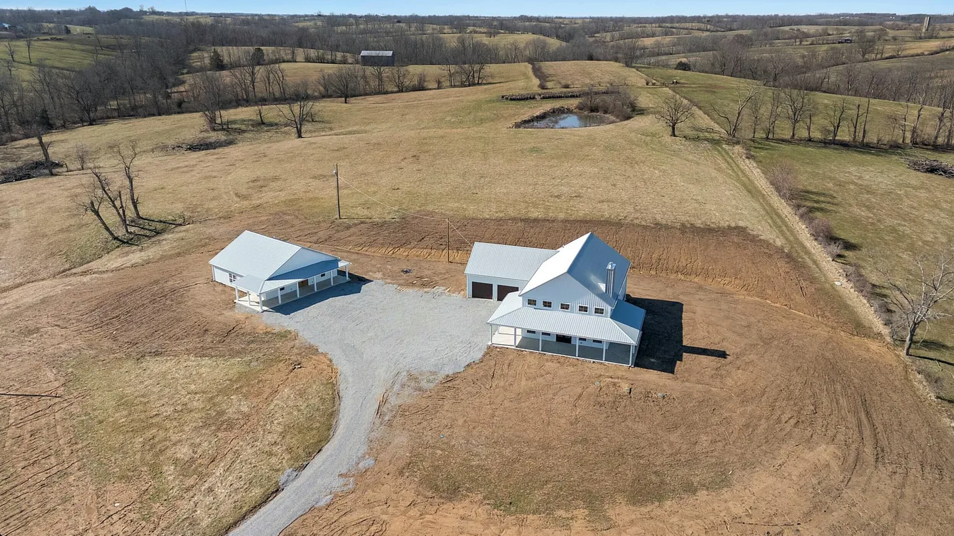 Aerial view of a white house and detached garage on a gravel driveway surrounded by large, grassy, rural fields.