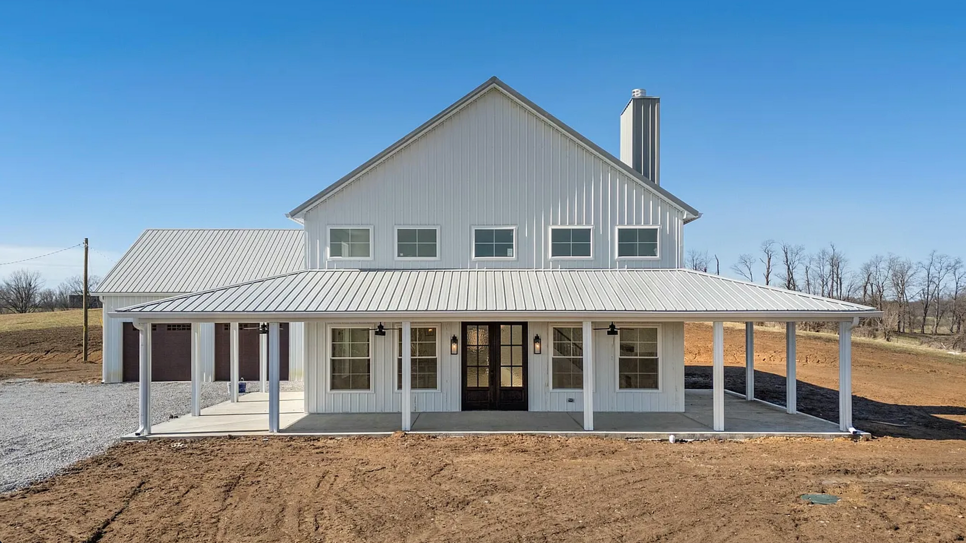 A white farmhouse with a large wraparound porch and a metal roof, set against a blue sky on a clear day.