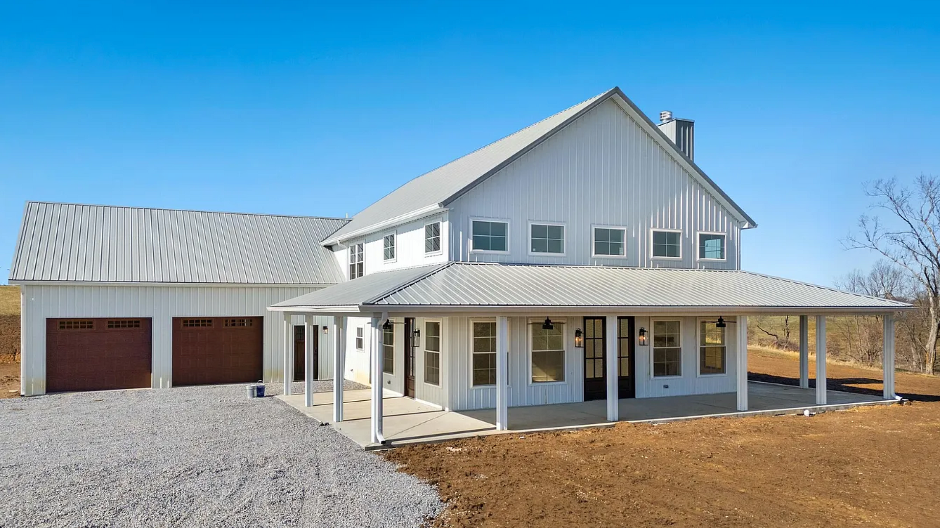 A two-story white farmhouse with a metal roof, a wrap-around porch, and attached garage doors under a clear blue sky.