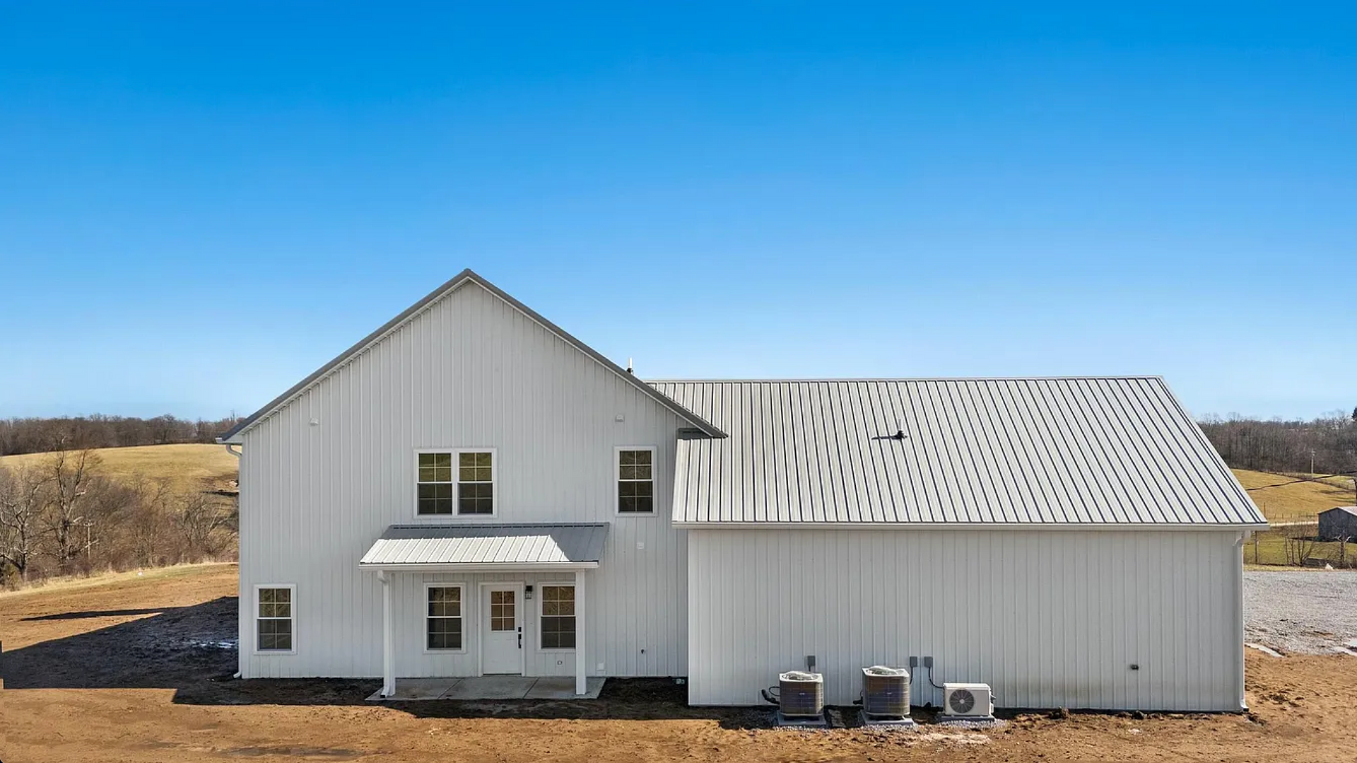 White two-story house with a metal roof and light siding sits in a vast, open field under a clear blue sky.