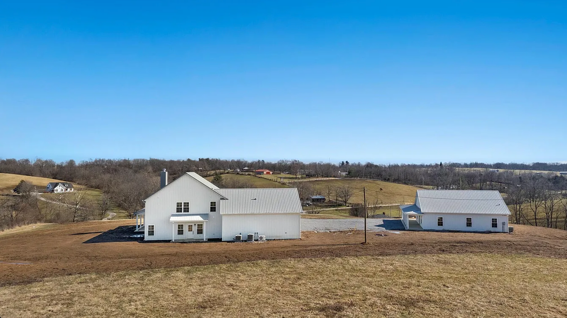 Two white farmhouse-style buildings with gray metal roofs sit on a grassy, rolling hill under a clear blue sky.