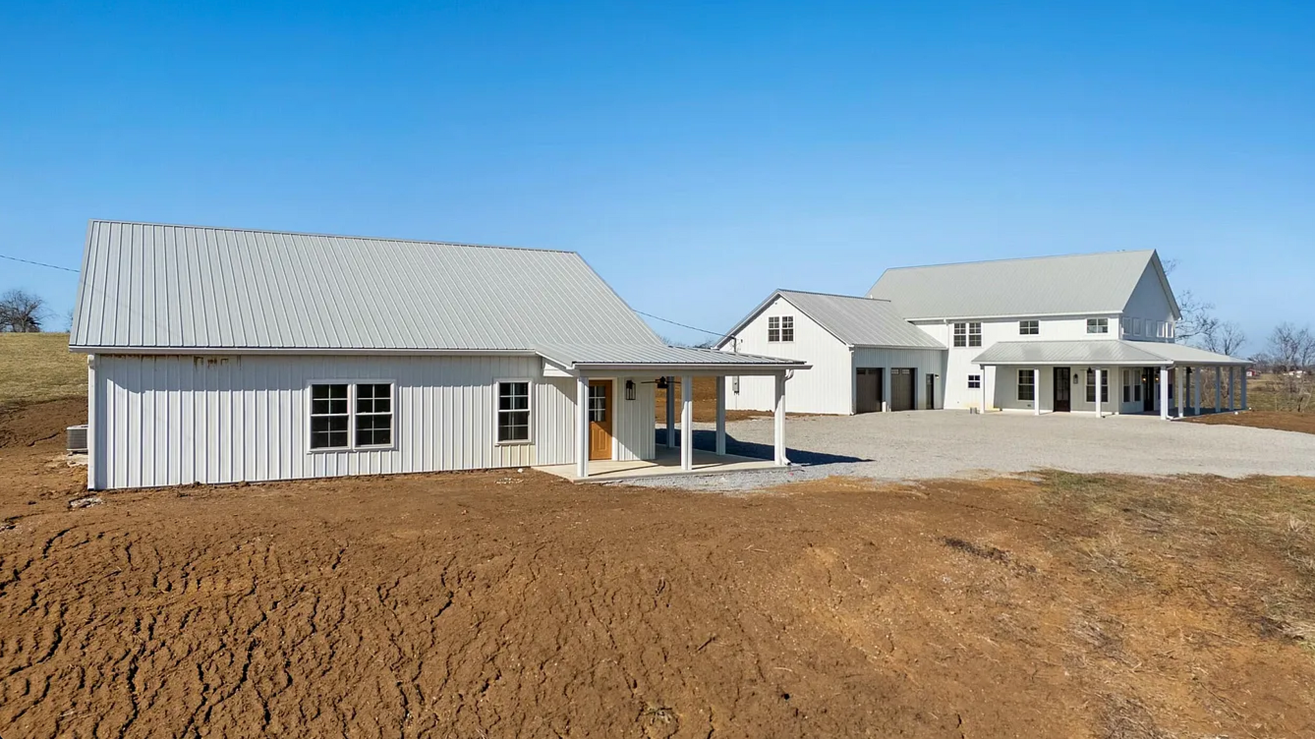 Two white buildings with metal roofs sit in an unlandscaped, dirt-covered lot under a clear blue sky.