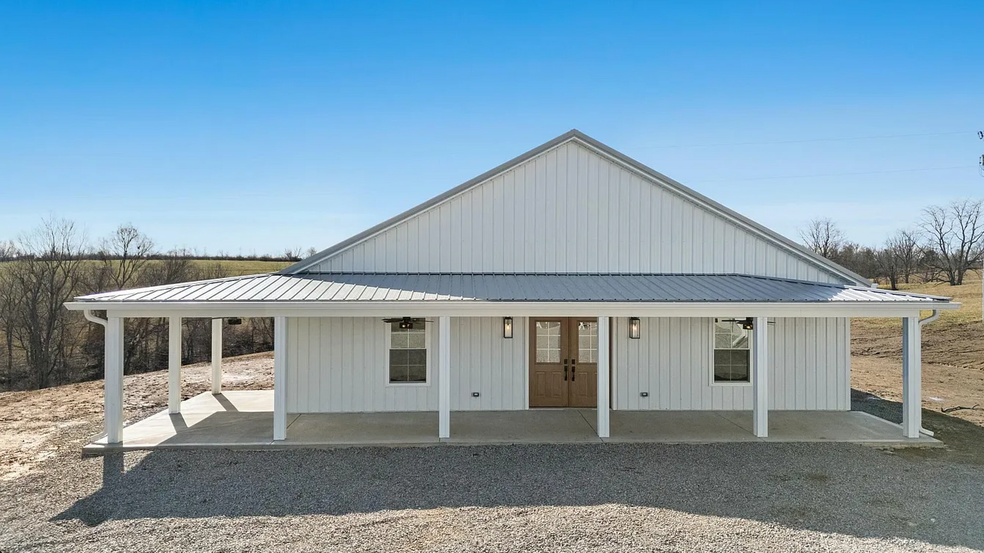 A white, metal-sided barndominium with a covered front porch, glass double doors, and a gravel yard under a clear blue sky.