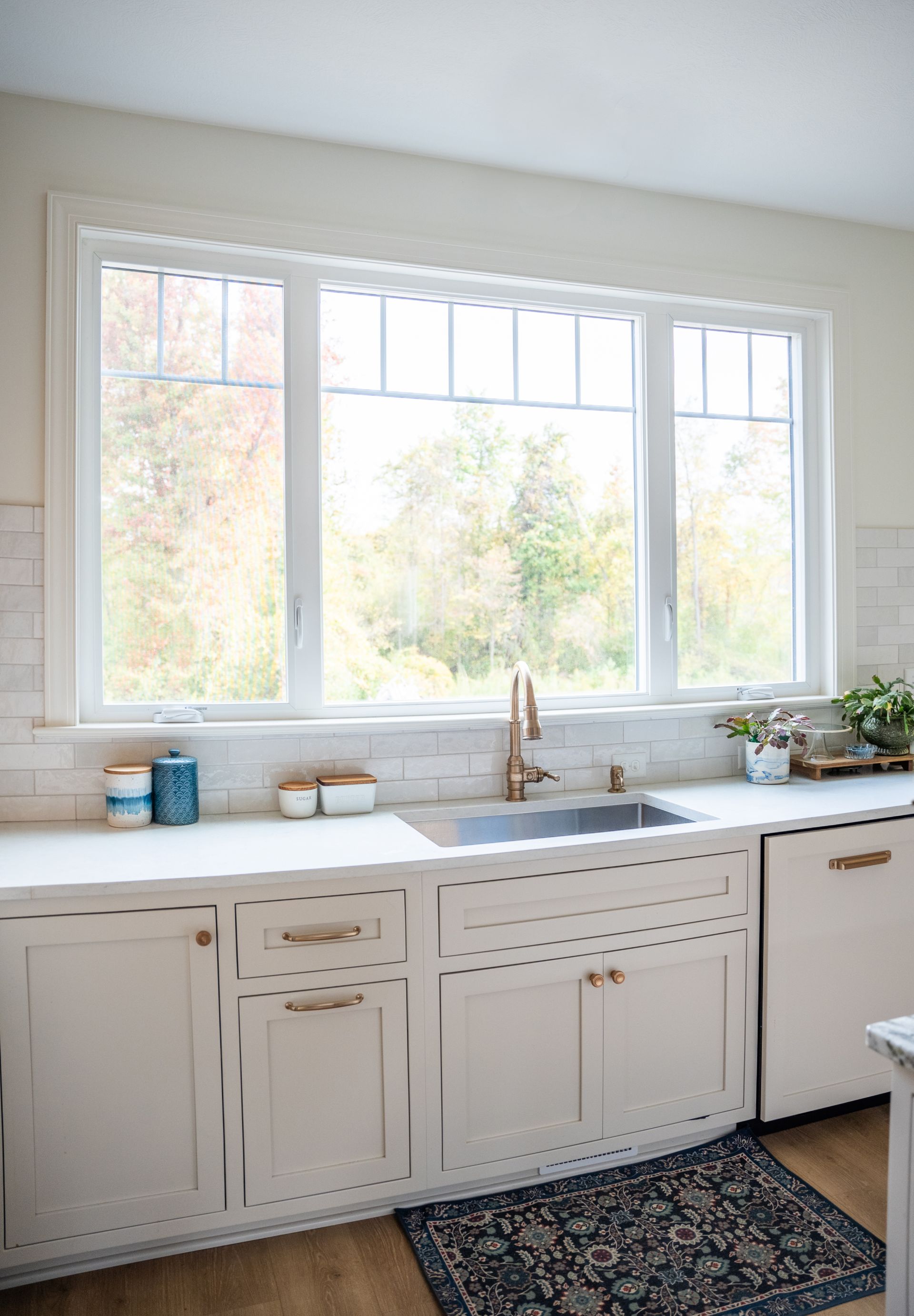 A bright kitchen sink area with light-colored cabinets, a stainless steel faucet, and a large window looking onto trees.