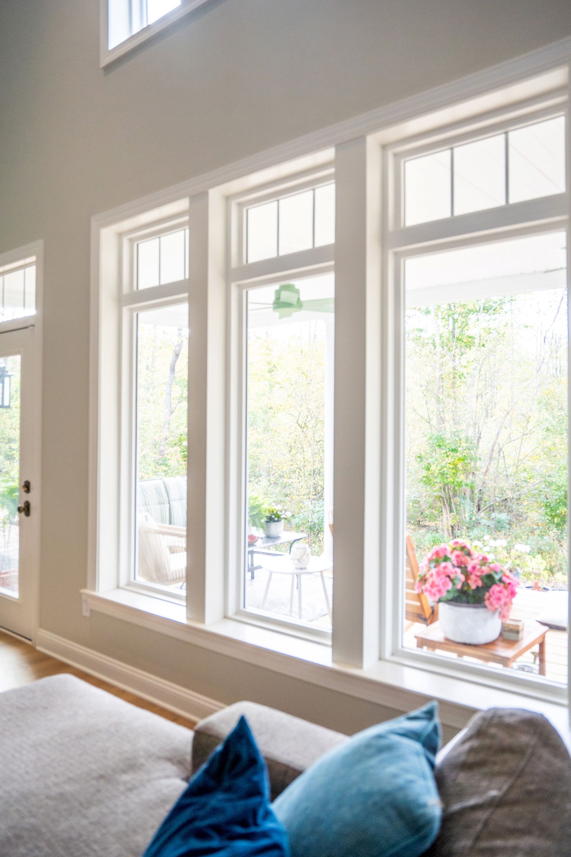 Tall, white-framed windows overlook a garden with greenery and a small pot of pink flowers, viewed from a cozy living room.