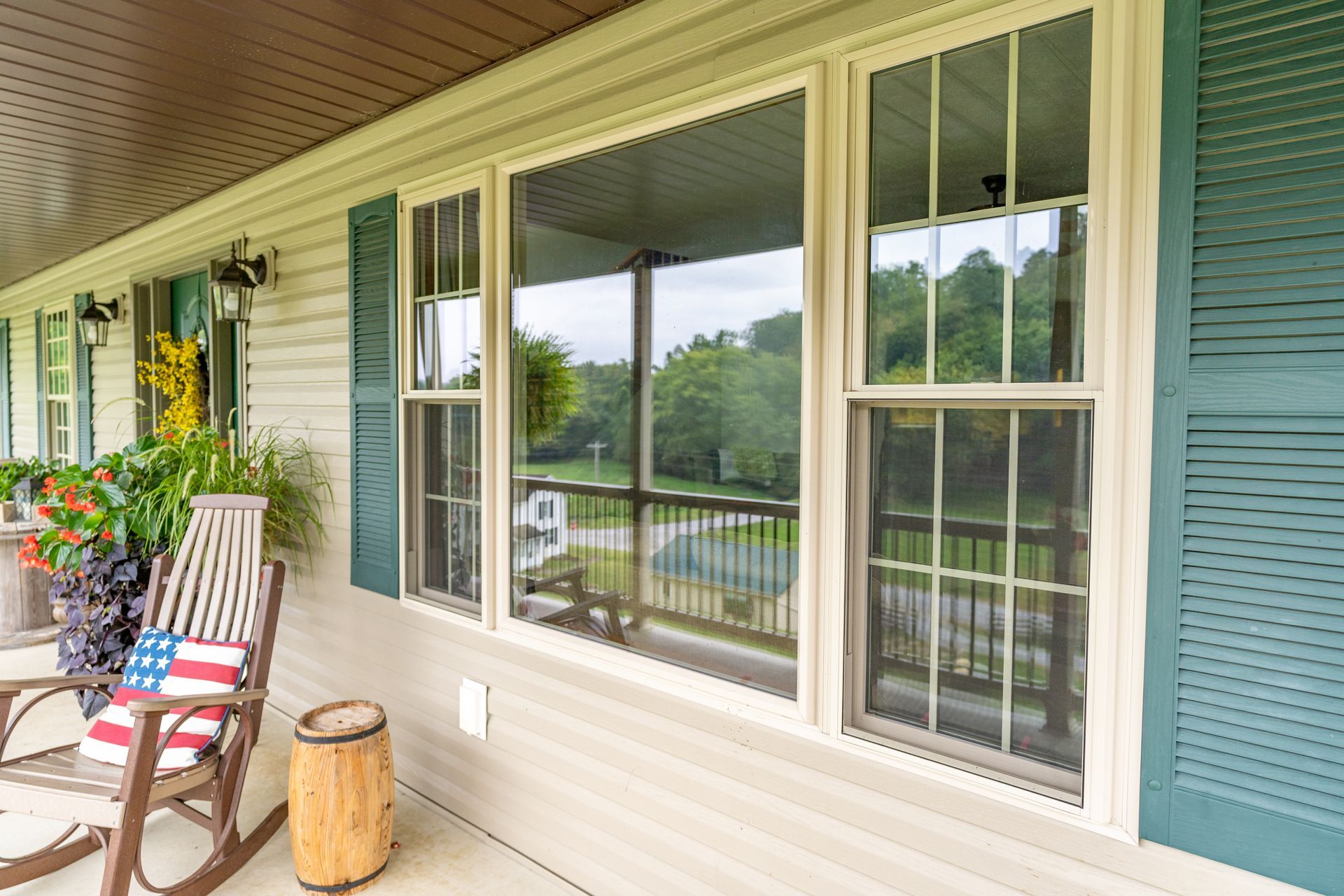 A front porch with a wooden rocking chair, a small barrel-shaped side table, and a window with green shutters.