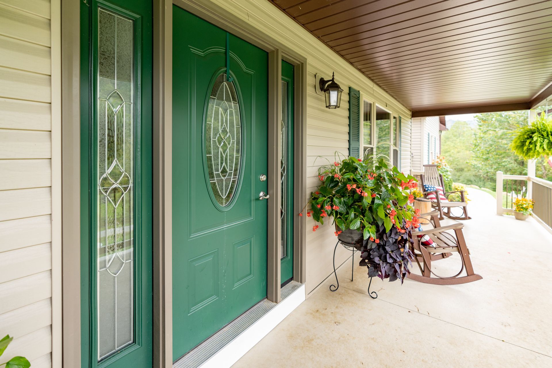 A green front door with a decorative glass oval on a porch featuring a large potted plant and two wooden rocking chairs.
