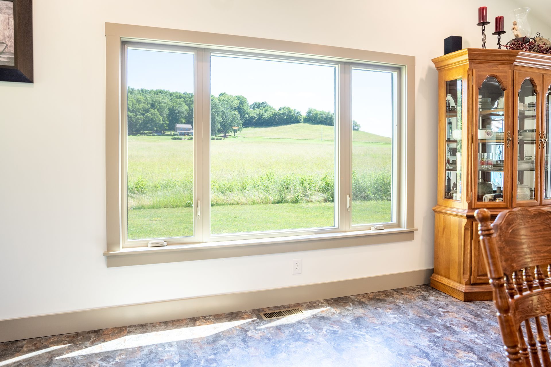 A wide, three-pane window framed in beige looks out onto a sunny, grassy hillside with trees.