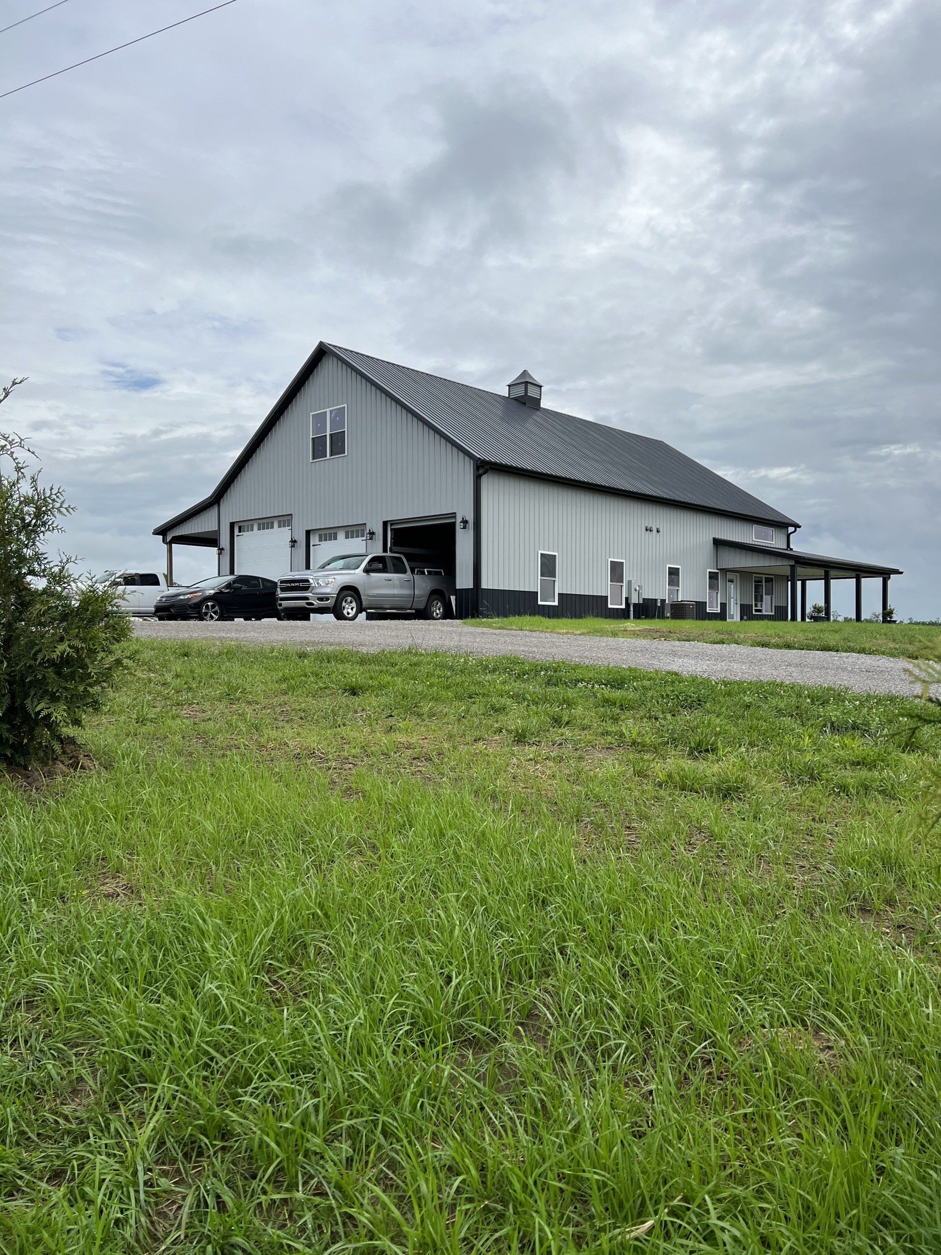 A large, white metal-sided barn-style building with a dark metal roof and a covered porch, set on a grassy hill.