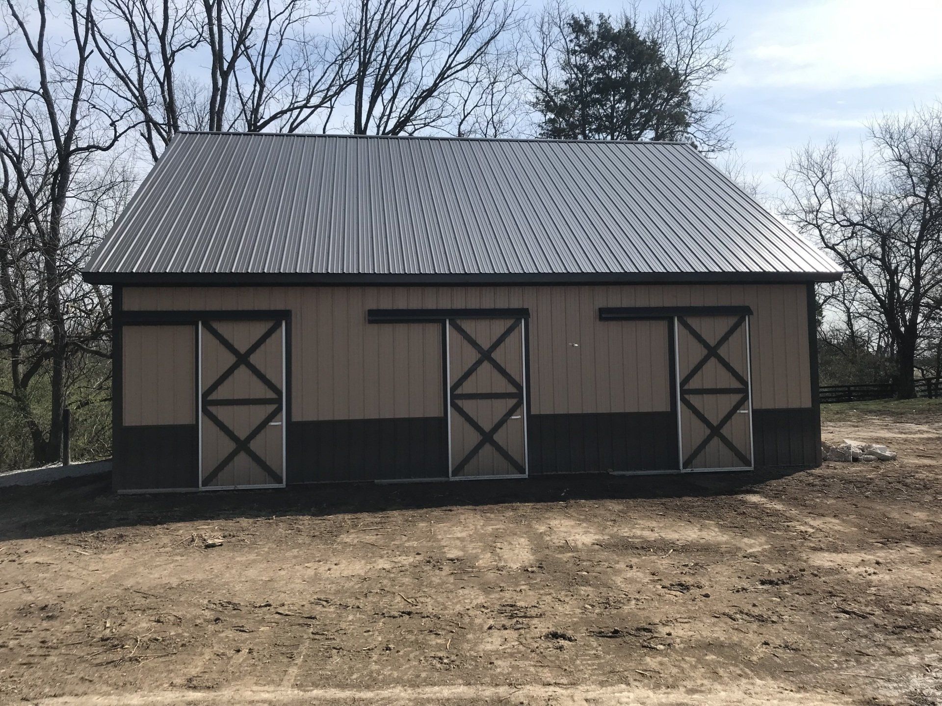 A tan and dark brown metal barn with three sliding doors and a gabled metal roof, situated on a dirt lot.