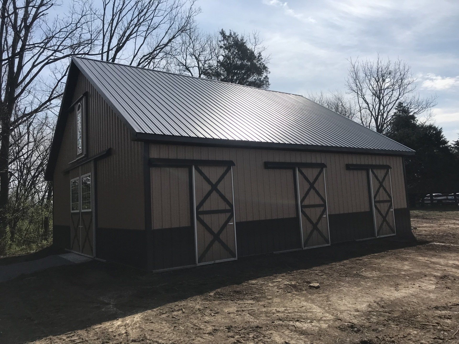A brown wooden horse barn with a dark metal roof and three sliding stall doors, situated on a rural dirt lot.
