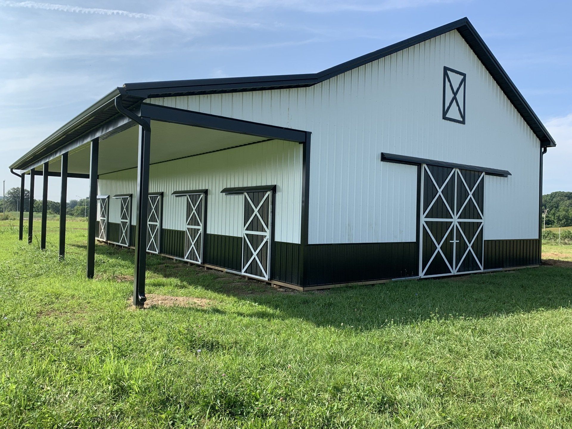 A white and black pole barn with a side porch, multiple stall doors, and a large sliding door in a grassy field.