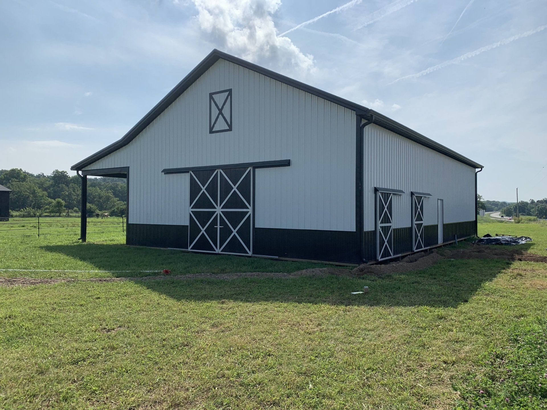 A white and black metal barn with sliding doors sits in a grassy field under a sunny sky.