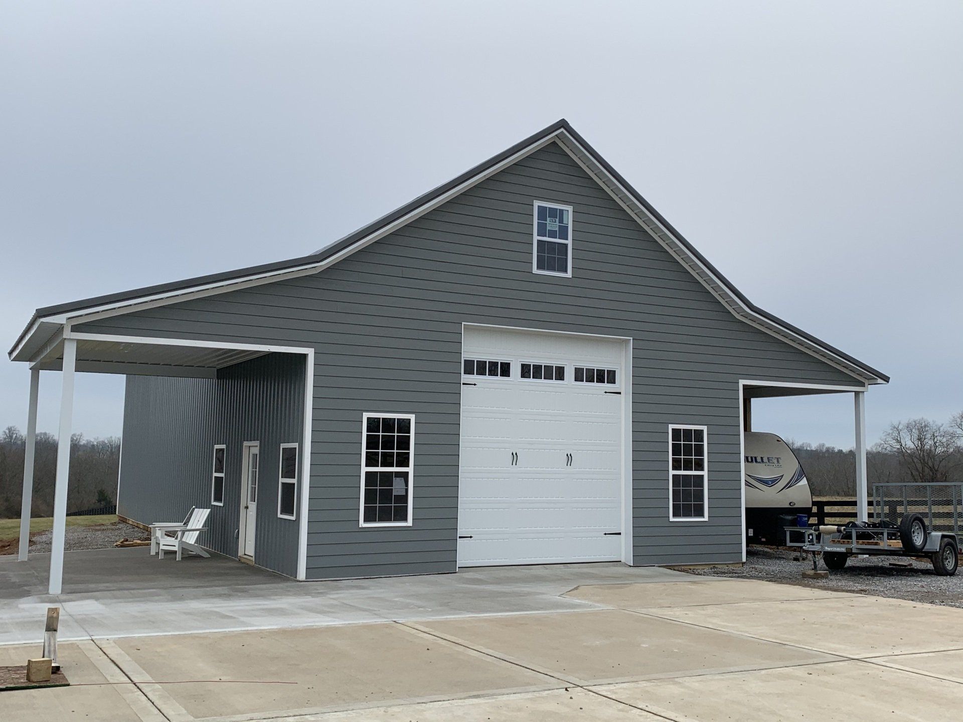 A grey metal-sided barn with a white garage door, two porches, and white-trimmed windows on a concrete lot.