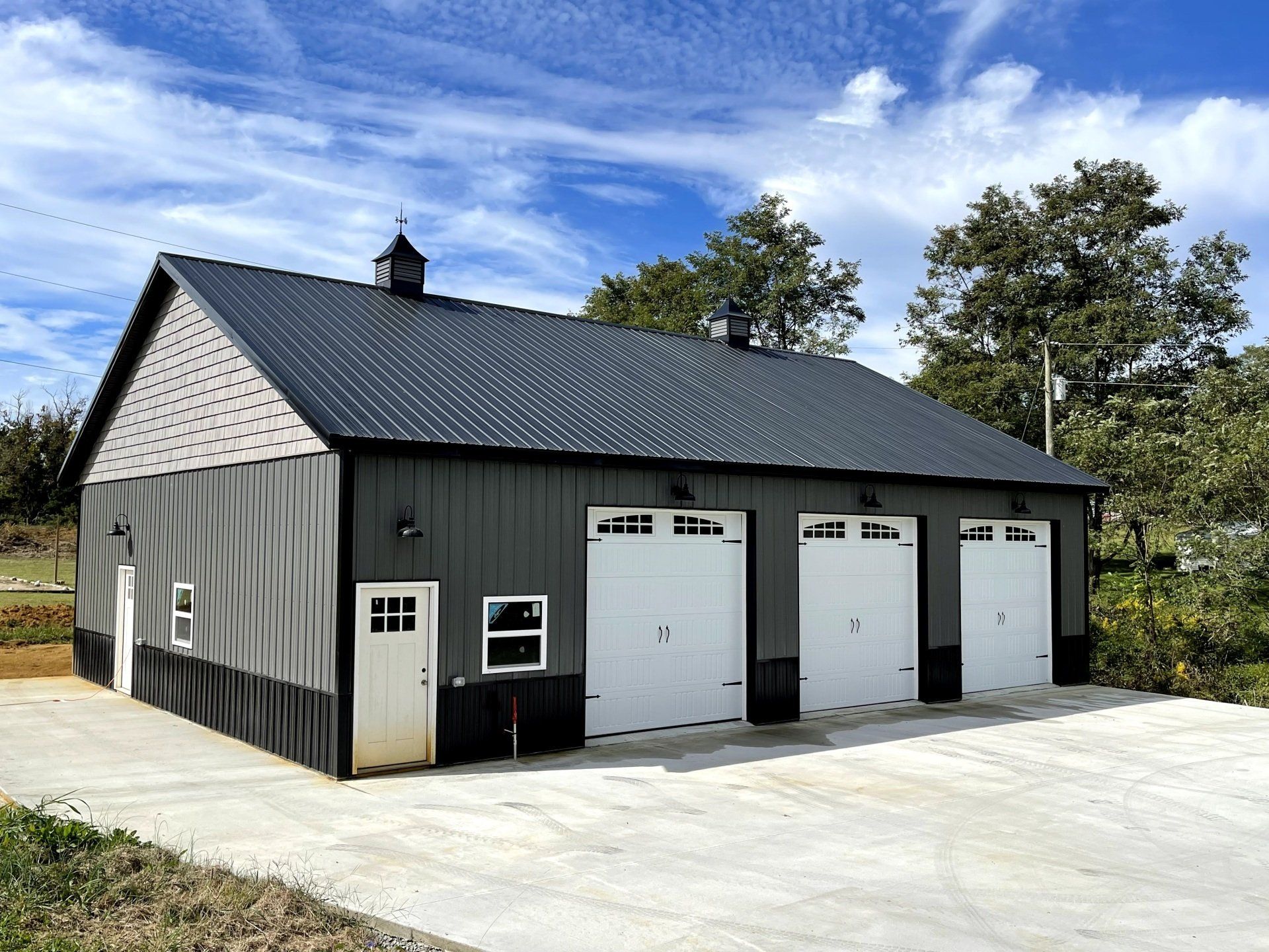A modern gray metal garage with a black roof, three white doors, and a small cupola on a paved concrete driveway.