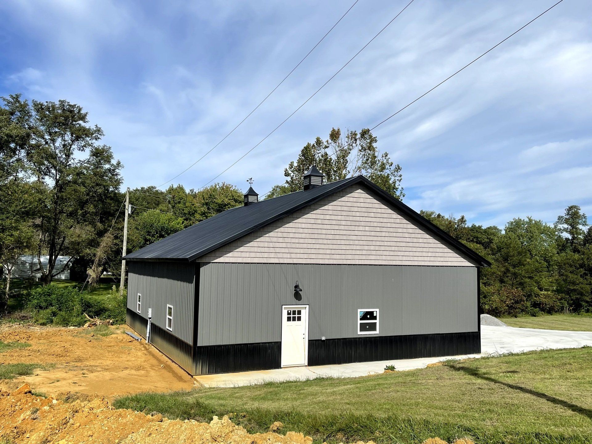 A gray metal barn with black trim, a white door, and two small cupolas on a black roof sits in a grassy field.