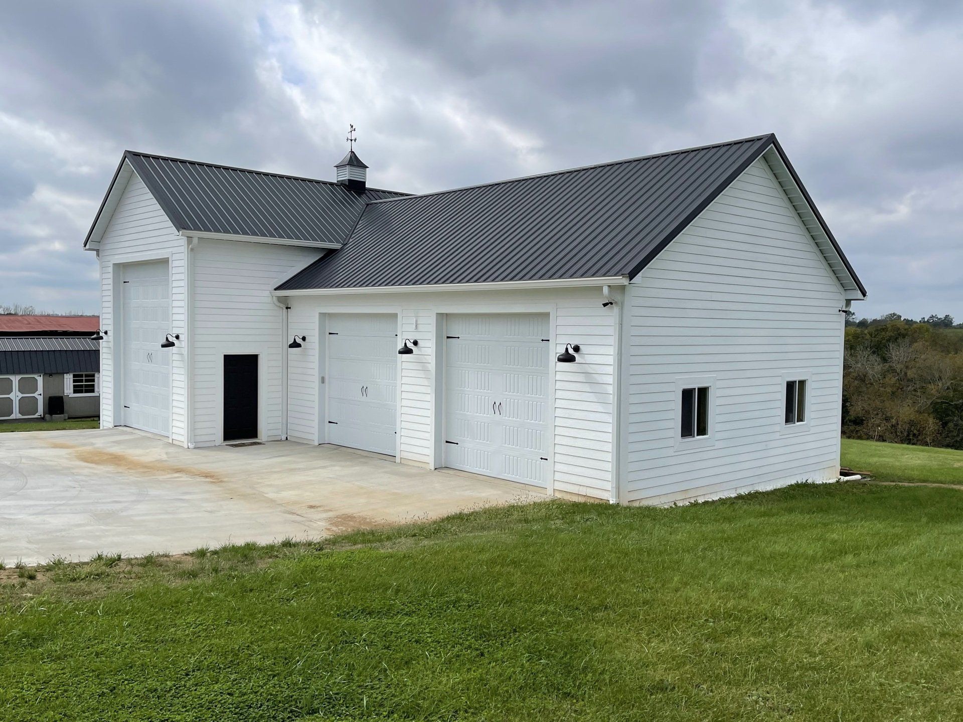 A white, multi-bay garage with a dark metal roof and black light fixtures, set on a grass lawn under a cloudy sky.