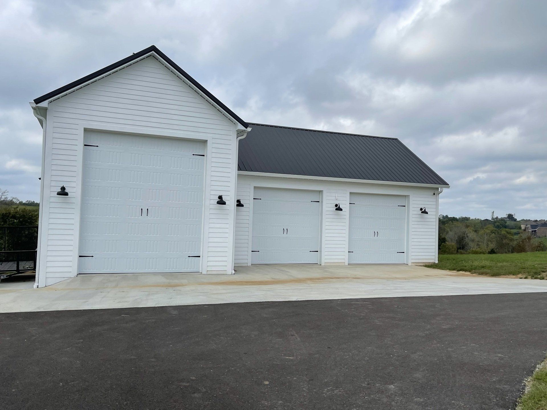 A white garage with three white bay doors and a dark metal roof, set against a cloudy sky with an asphalt driveway.