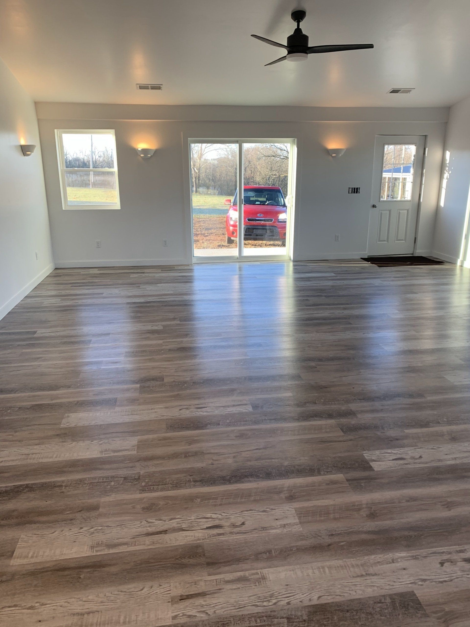 A spacious room with wood-look flooring, white walls, wall sconces, a ceiling fan, a sliding glass door, and a white door.