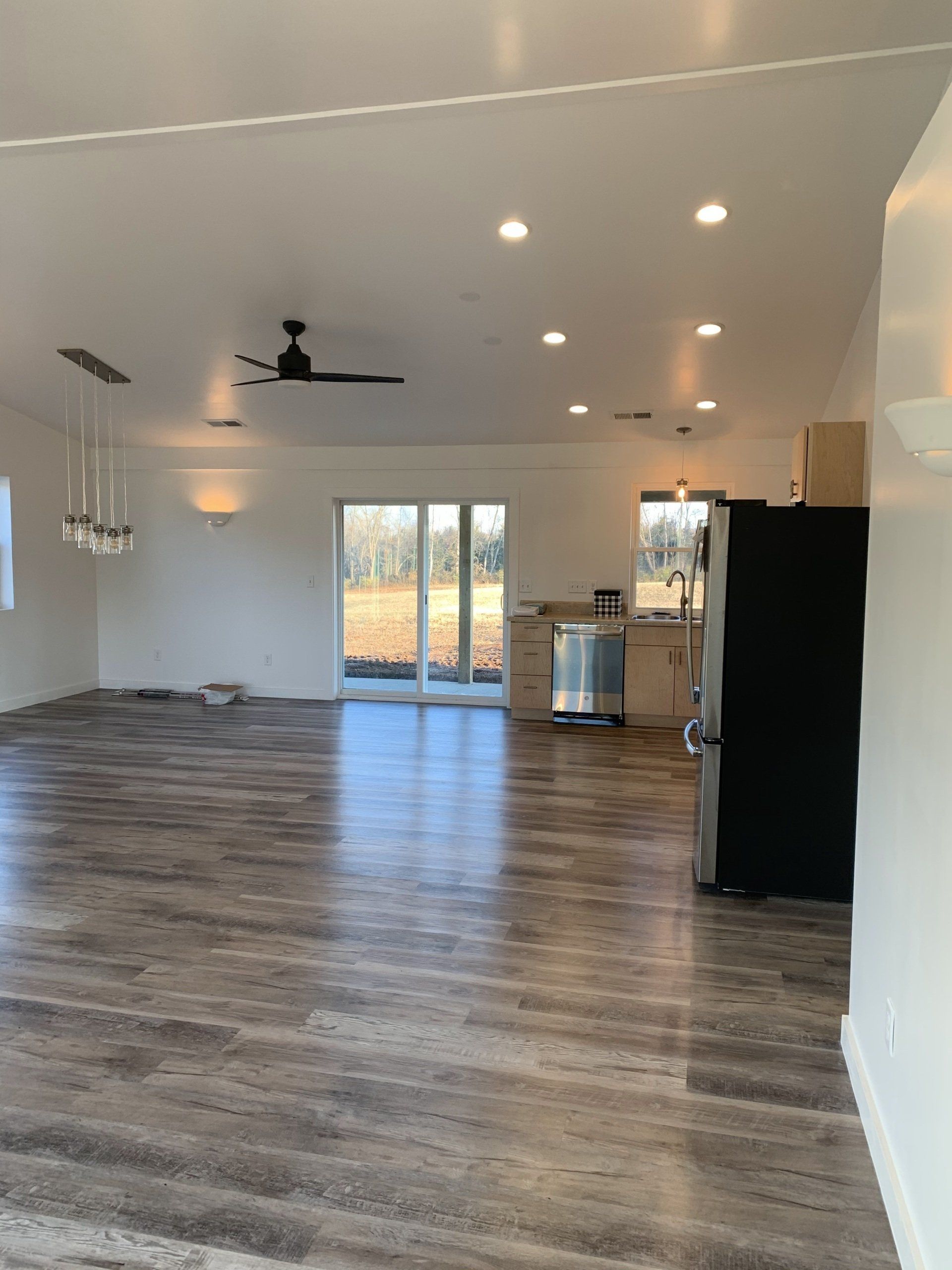 A spacious, modern open-concept room with grey wood-look flooring, recessed lighting, a ceiling fan, and a kitchen area.