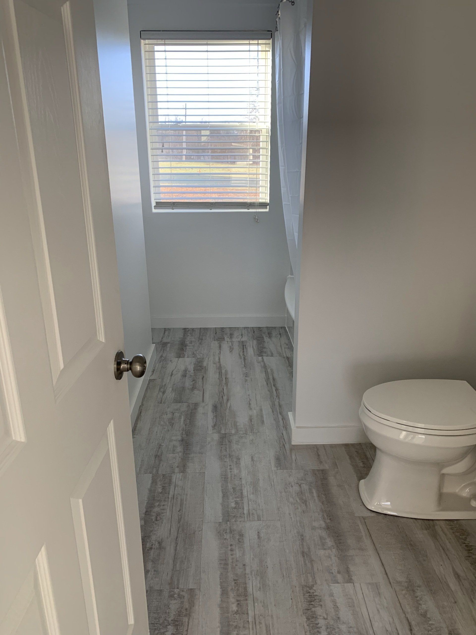 A view of a bathroom doorway, showing grey wood-look flooring, a toilet on the right, and a window with blinds at the back.