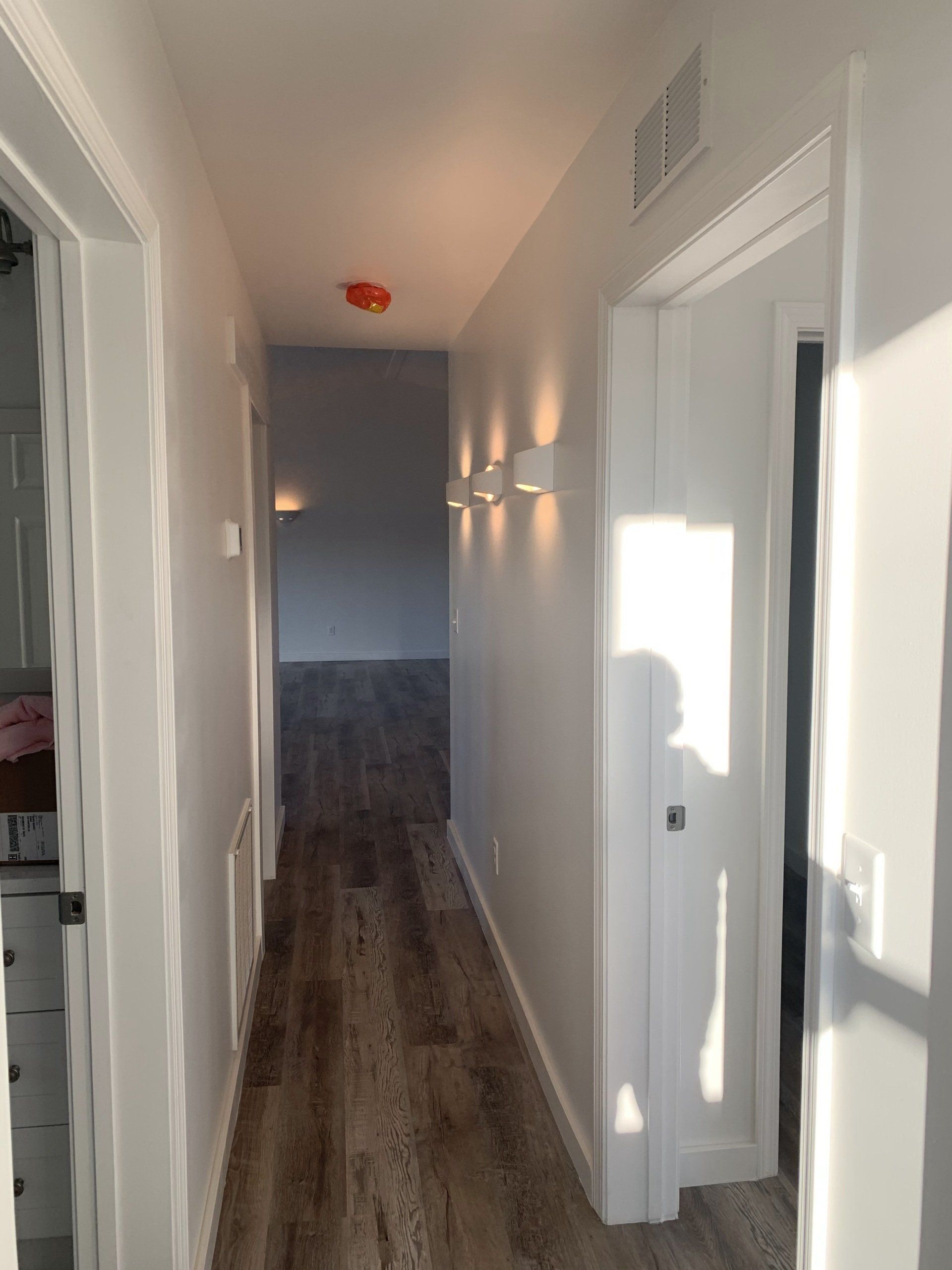 A narrow, wood-floored hallway with white walls, illuminated by three modern wall sconces and a ceiling light.