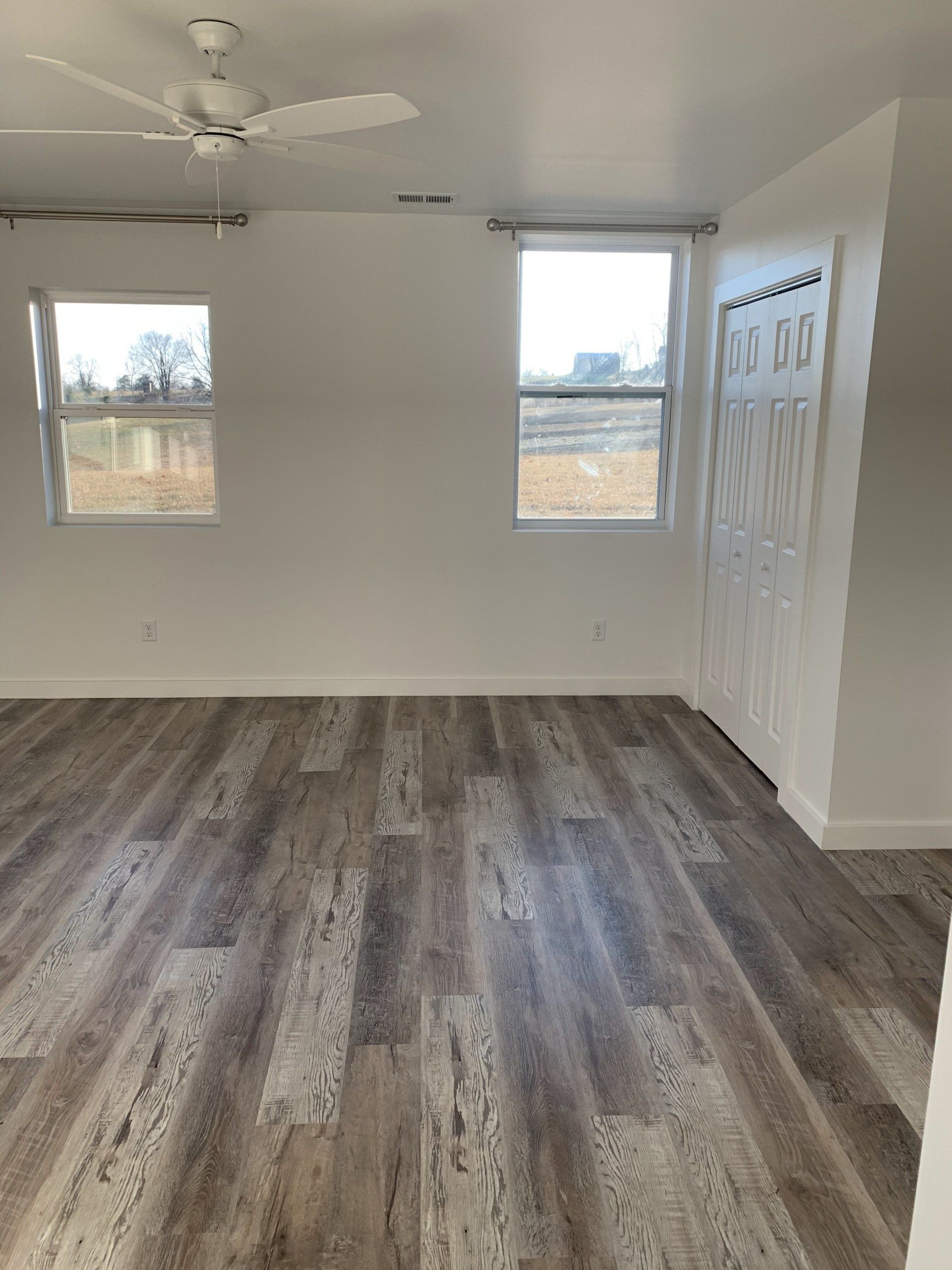 An empty room with light gray wood-look flooring, white walls, two windows overlooking a field, and a ceiling fan.