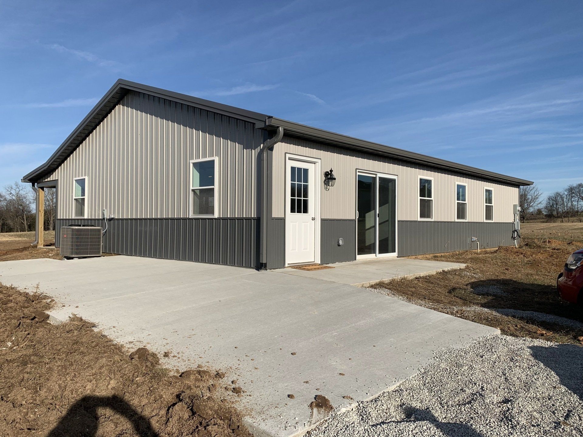 A modern metal-sided house with a dual-tone gray exterior, white doors, and a concrete patio on a sunny, rural lot.