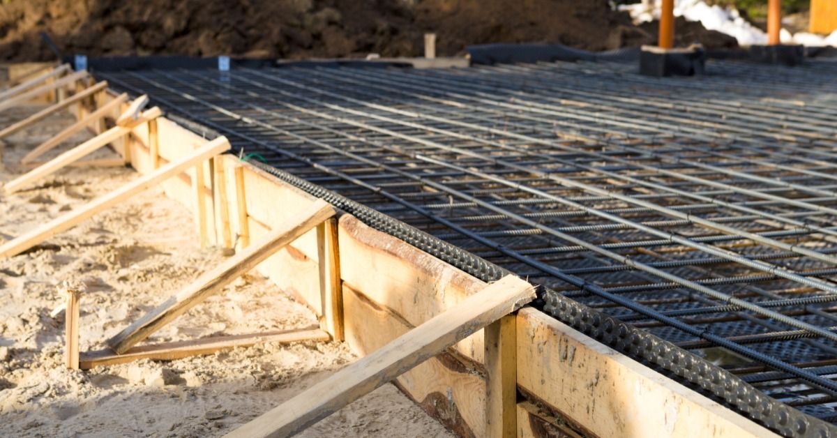 A wooden formwork with metal reinforcement bars and laid pipes arranged in preparation for pouring a concrete foundation slab.