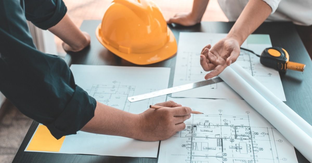 A group of engineers and architects gathered around a table reviewing blueprints with a laptop, rulers, and measuring tools.