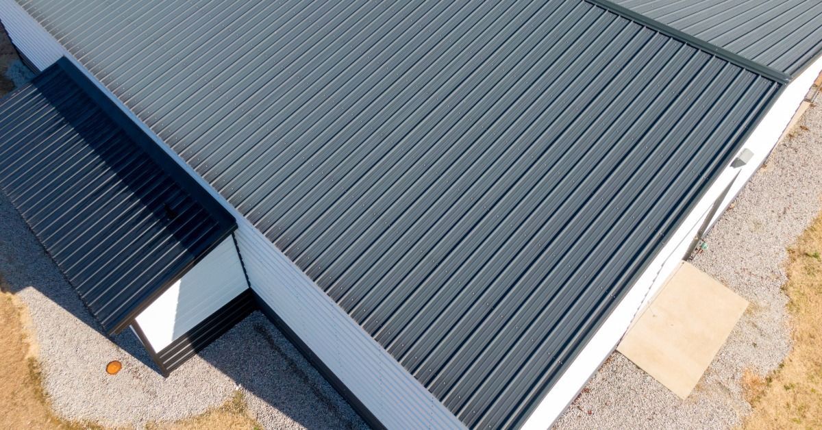 An aerial view of a gray metal roof with vertical seams, vents, and a light-colored building edge.