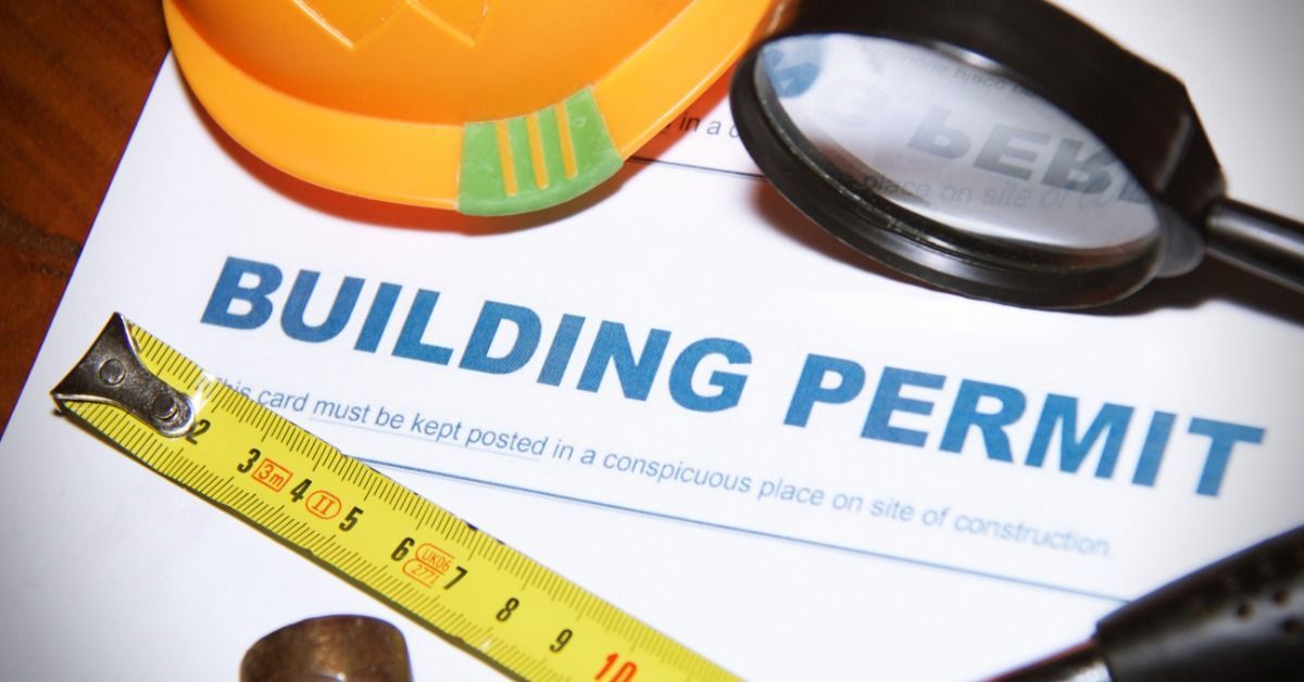 A building permit form with a hard hat, magnifying glass, tape measure, stamp, and pen on a wooden table.