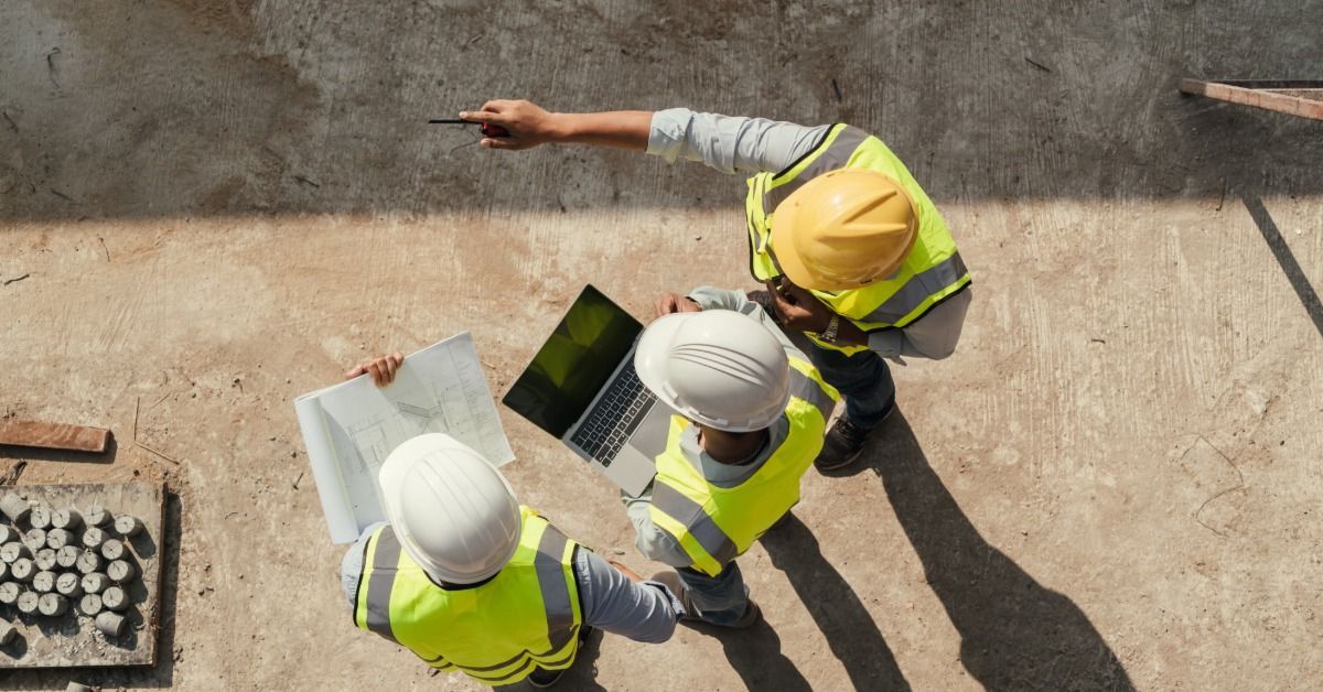 Three people in hard hats reviewing a blueprint and laptop on a concrete surface at a construction s
