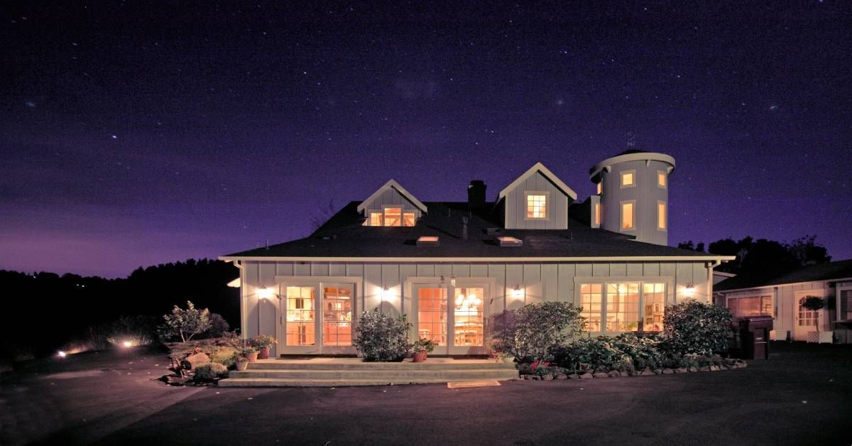 A white barn house lit up with warm indoor and outdoor lighting. Above the house is a blue-black starry sky.