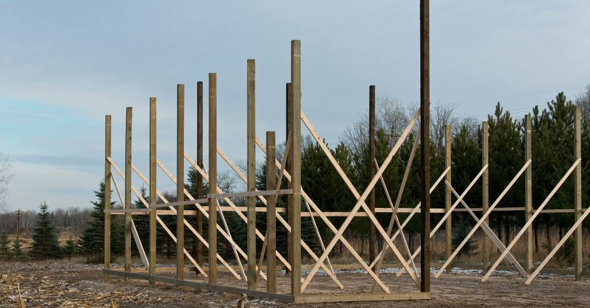 The basic wooden frame of a pole barn under construction in a field surrounded by dirt and dead cornstalks.