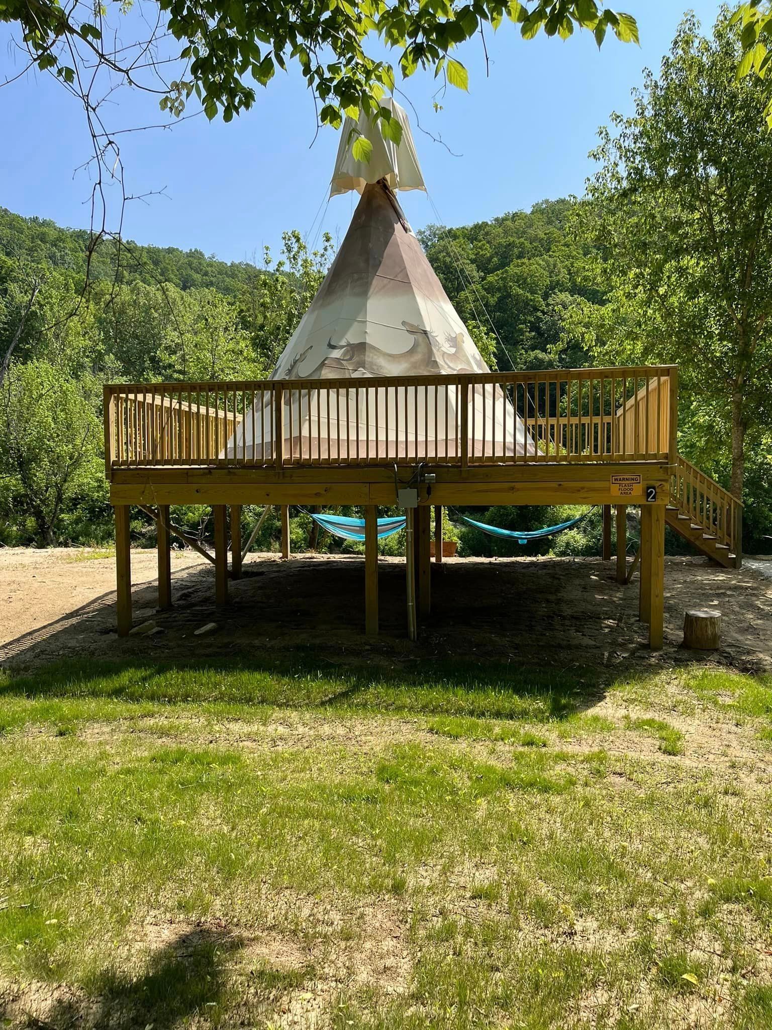 A teepee structure with a weathered white cover sits on a wooden deck in a grassy, wooded outdoor setting under blue skies.