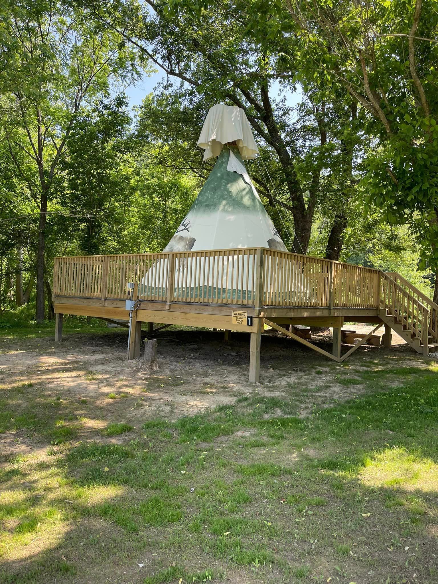 A teepee tent sits on a raised wooden deck surrounded by trees in a grassy, sunlit outdoor area.