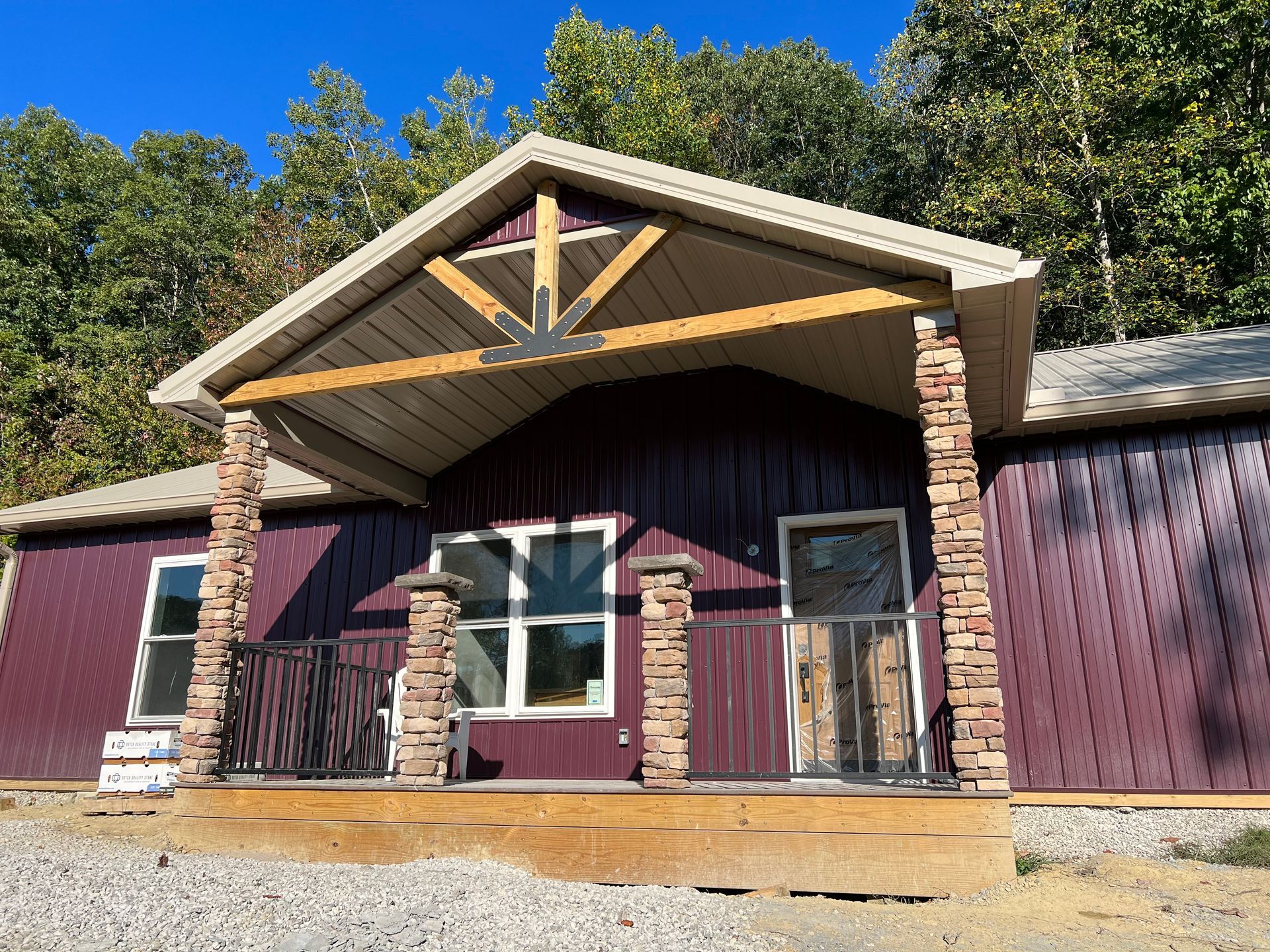 A maroon cabin with a stone-pillared porch, a gabled roof, and a gravel yard, set against a backdrop of green trees.