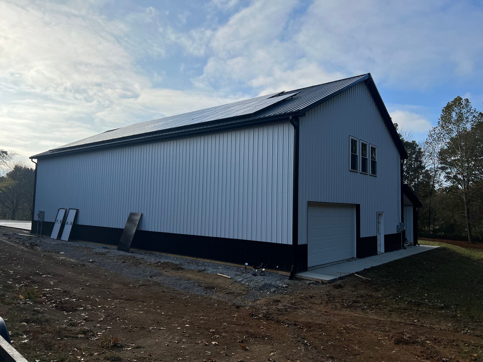 A large metal building with white vertical siding and black trim sits on a graveled lot under a cloudy blue sky.