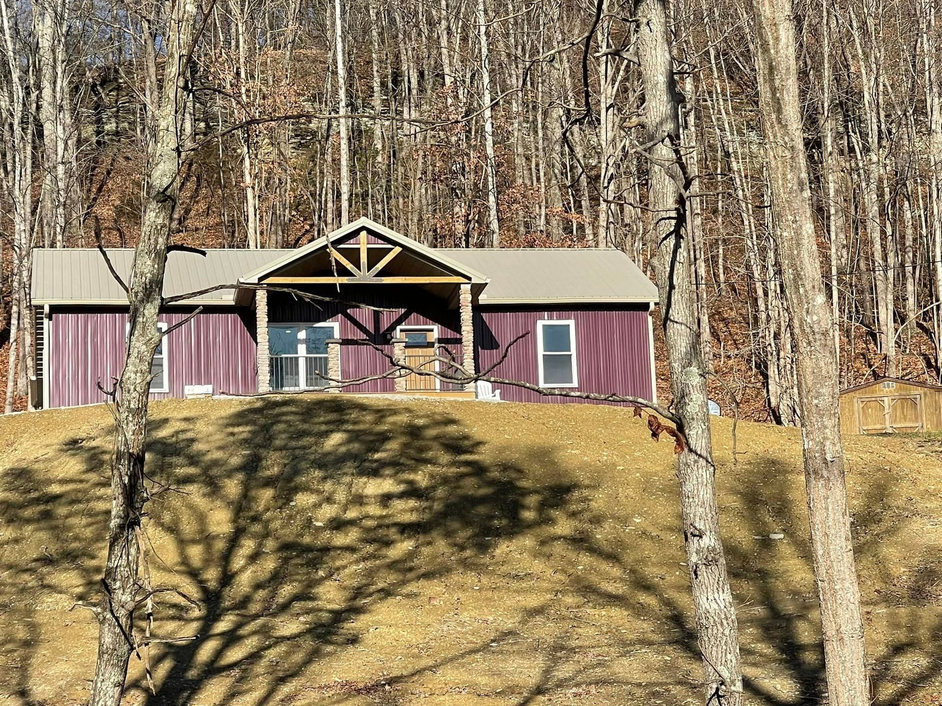 A purple, single-story cabin with a metal roof and a wooden front porch, situated on a grassy hill in a wooded area.