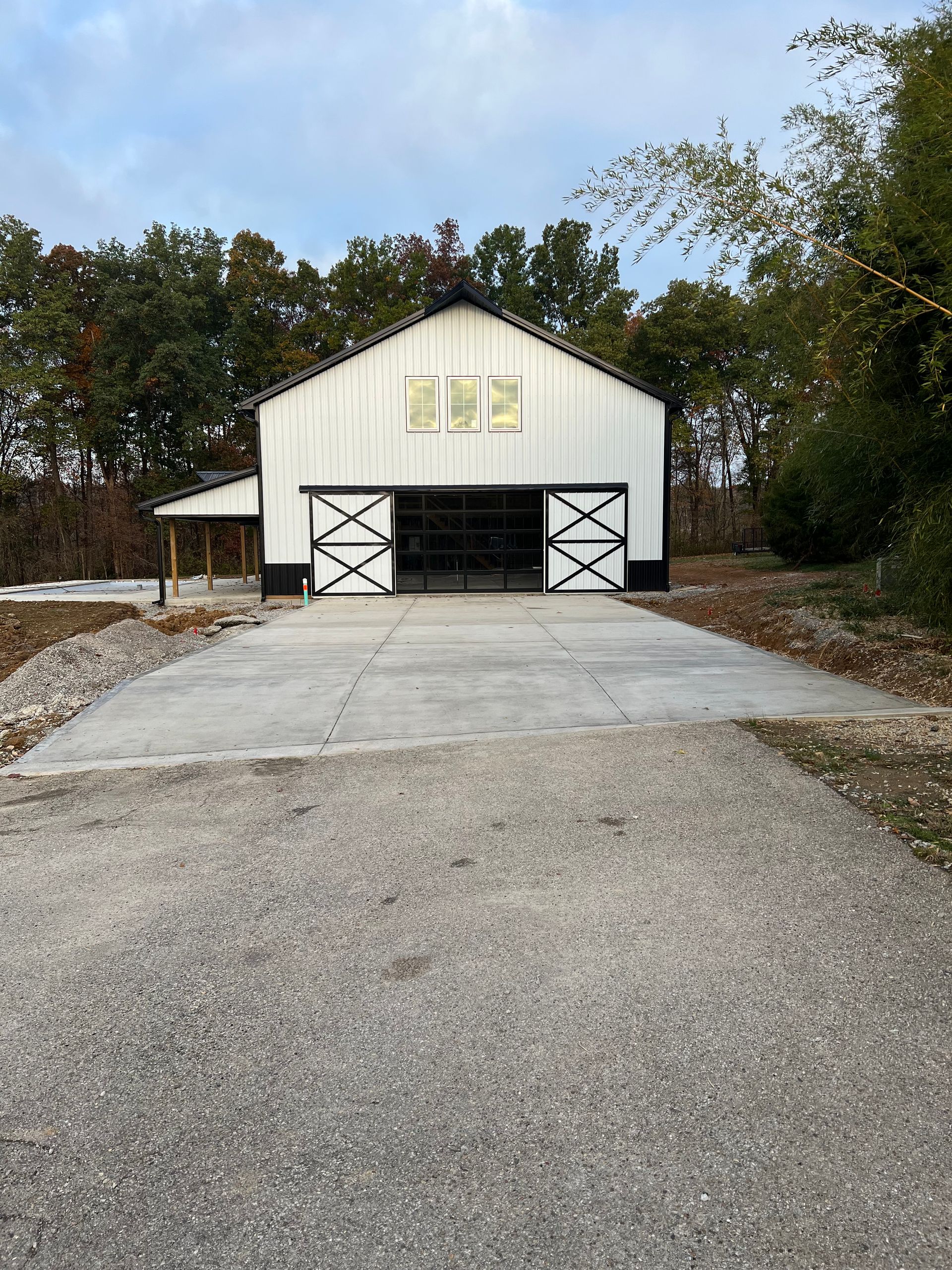 A white metal barn with black trim and sliding barn-style doors, situated at the end of a gravel and concrete driveway.