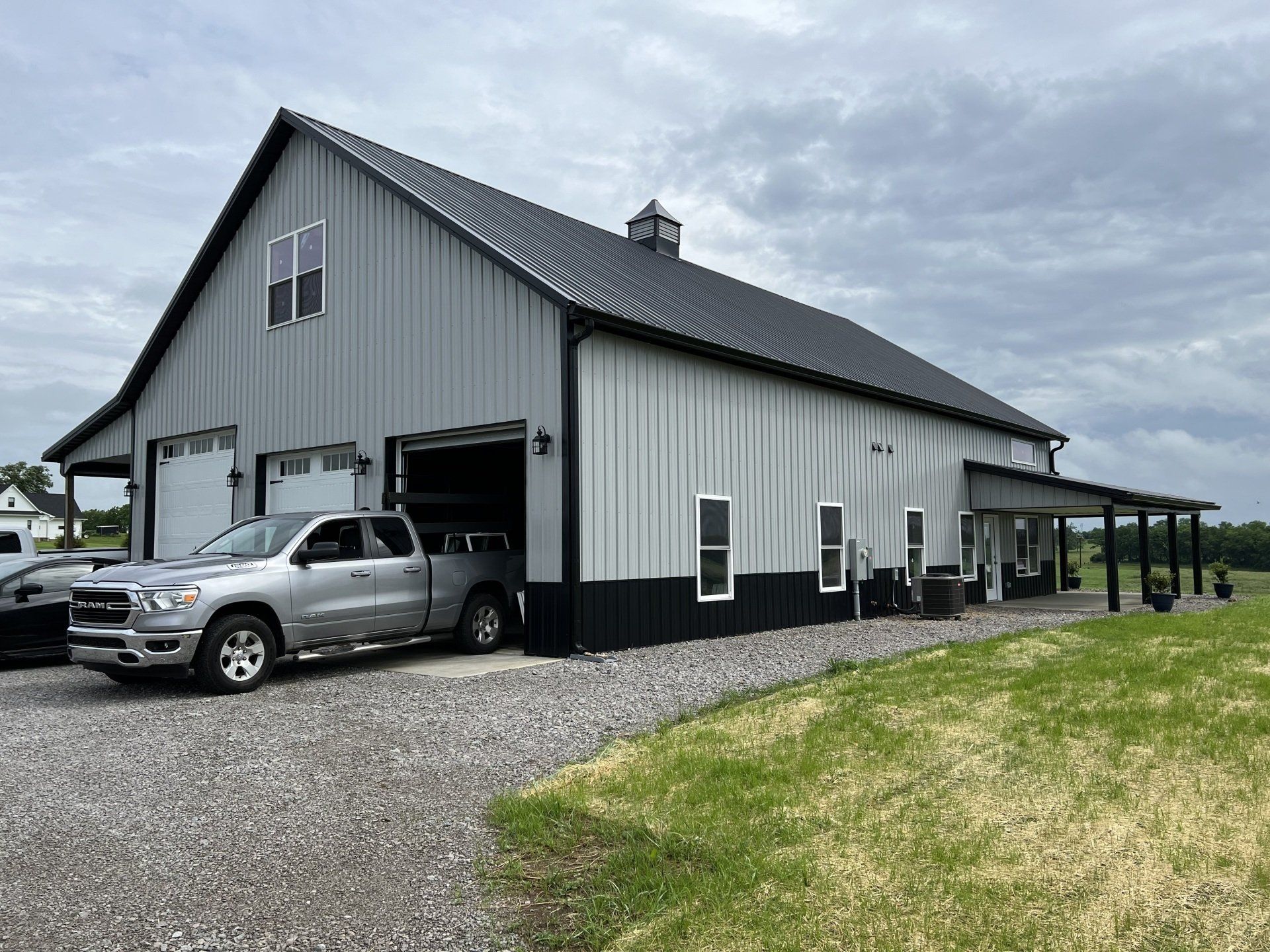 A grey and black metal barndominium with a parked silver pickup truck on a gravel driveway under a cloudy sky.