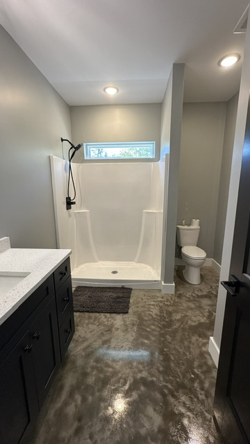 A modern bathroom with a white walk-in shower, a dark vanity, a toilet in a nook, and polished concrete flooring.