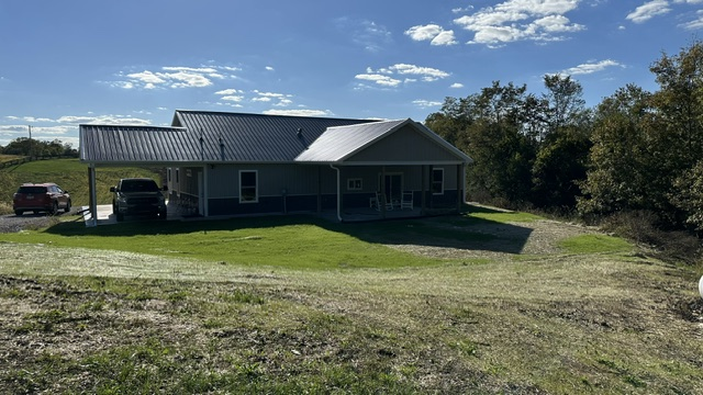 A tan house with a metal roof and covered porch stands on a grassy lawn under a blue sky with scattered clouds.