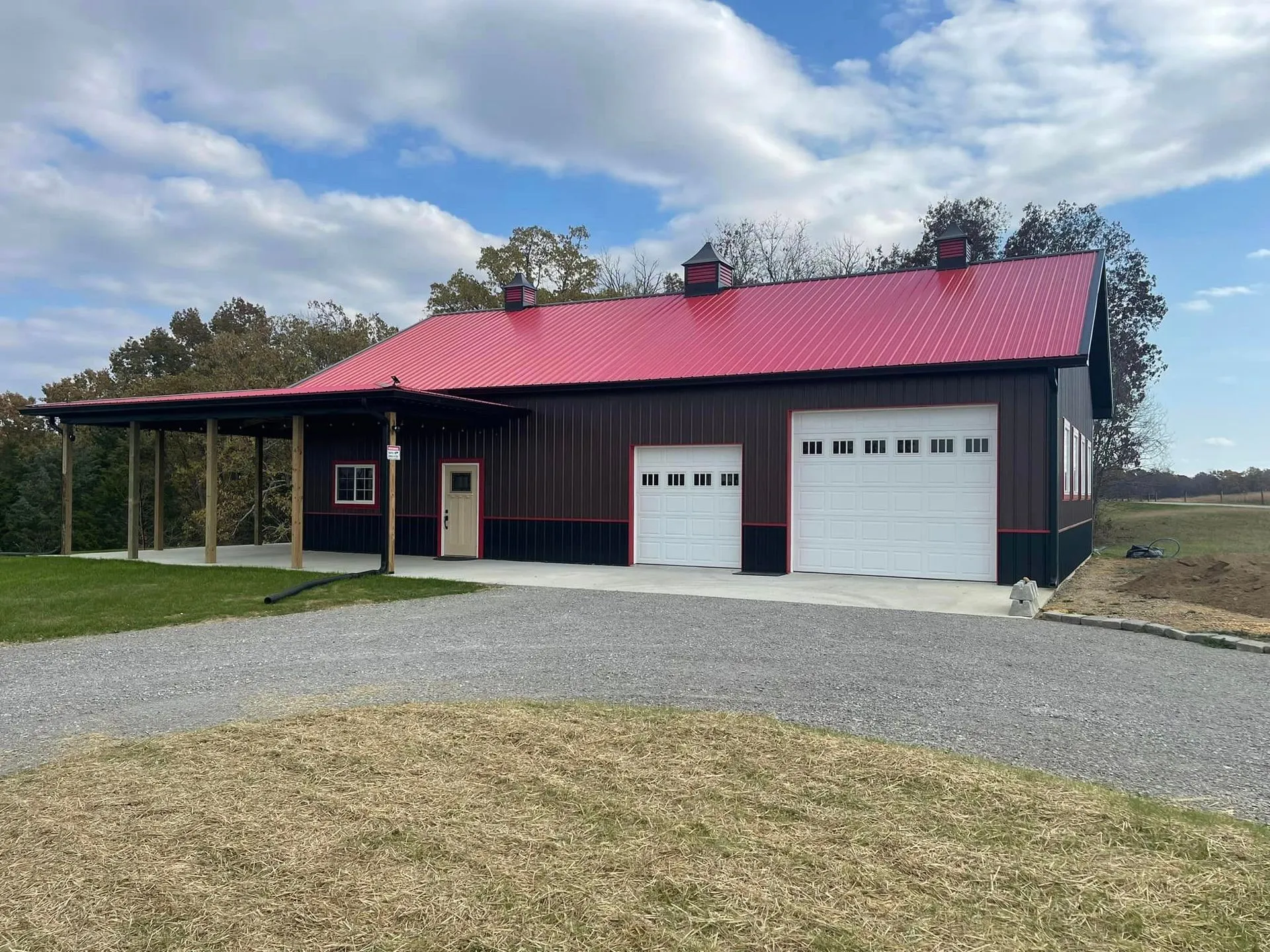 A modern metal building with a bright red roof, dark walls, a front porch, and two white garage doors, set near trees.